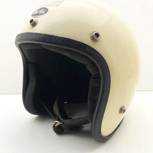 Peut inclure: Casque de moto crème brillant. Le casque a une bordure noire autour de l'ouverture et un intérieur noir. Un petit logo est visible sur le côté. Comprend une jugulaire.