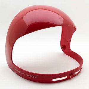 Può includere: Visiera per casco rossa lucida, curva e aerodinamica, con una parte superiore arrotondata e una fascia inferiore. La fascia presenta ritagli e la scritta "DESIGN BY F.A. PORSCHE". Realizzata in materiale duro e resistente.