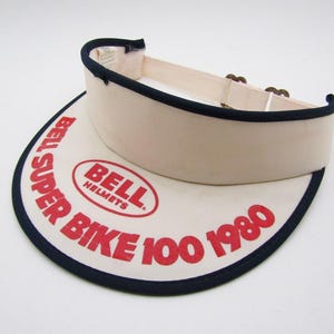 Può includere: Visiera color crema con bordatura blu navy e la scritta "BELL SUPER BIKE 100 1980" in rosso. La visiera ha una tesa curva e una fascia in tessuto con chiusure regolabili.