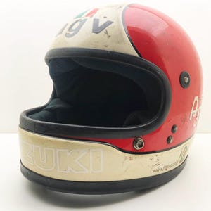 Può includere: Casco da moto vintage rosso e crema con la scritta "SUZUKI" in bianco. Il casco ha interni e finiture neri, con le lettere "IGV" e la bandiera italiana sulla parte superiore. Realizzato in fibra di vetro.