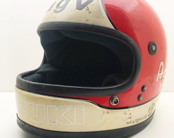 May include: Vintage red and cream-colored motorcycle helmet with the word "SUZUKI" in white lettering. The helmet has a black interior and trim, with the letters "IGV" and the Italian flag on top. The helmet is made of fiberglass.
