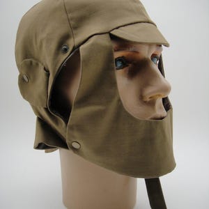 May include: A brown,  canvas winter hat with a flap that covers the face. The hat has a brim and a chin strap.