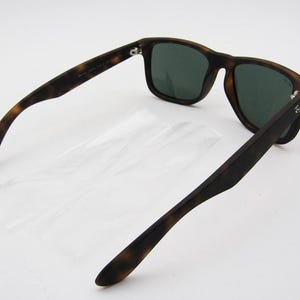 Puede incluir: Gafas de sol con estampado de carey y lentes verde oscuro. Las gafas tienen una montura rectangular cl&aacute;sica. Las patillas est&aacute;n abiertas, revelando el interior. El logotipo de Ray-Ban es visible en la patilla.