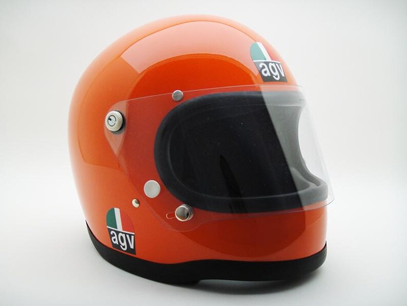 May include: Glossy orange motorcycle helmet with a clear visor and black interior. Features the "agv" logo in white on a green, white, and red background. The helmet has a black base and silver hardware.