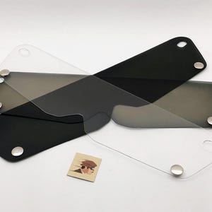 Peut inclure: Trois visières de casque de moto en clair, teinté et noir. Chaque visière a une forme incurvée avec des coins arrondis et des fixations métalliques. Une petite carte avec un graphique d'une personne portant un chapeau est également présente.