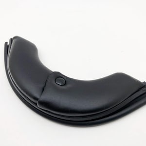 Può includere: Oggetto curvo in pelle nera con un dettaglio a bottone. L'articolo sembra essere una copertura protettiva o un rivestimento, forse per un mobile o un'attrezzatura. Il design è elegante e moderno.