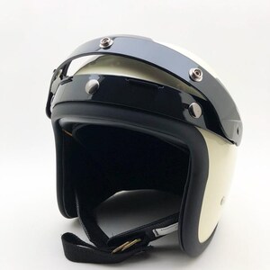 Puede incluir: Un casco de moto abierto de color blanco roto con una visera negra brillante sujeta por tres botones de presión metálicos plateados. El casco tiene un borde negro mate a lo largo de la abertura y una correa negra con hebilla metálica.