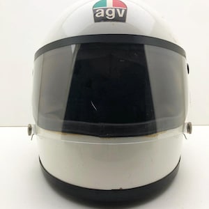 May include: White motorcycle helmet with a dark visor. The helmet has a black trim and a small emblem with the letters "agv" and a green, white, and red design. The helmet is designed for safety and protection.