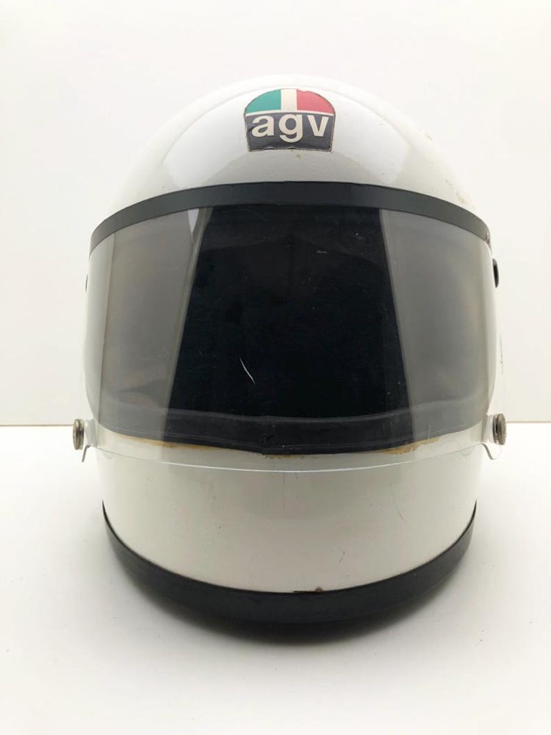 May include: White motorcycle helmet with a dark visor. The helmet has a black trim and a small emblem with the letters "agv" and a green, white, and red design. The helmet is designed for safety and protection.