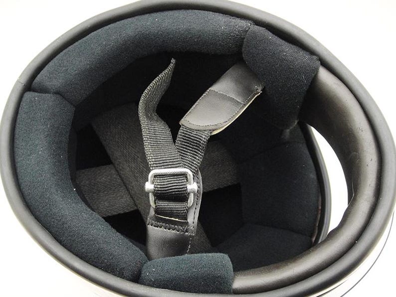 May include: Interior view of a motorcycle helmet. The helmet features a black padded interior with a chin strap and a metal buckle. The outer shell is white with a black trim.