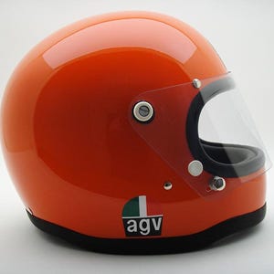 May include: Glossy orange motorcycle helmet with a clear visor and black trim. The helmet has a small green, white, and black logo on the side. A classic design for motorcycle enthusiasts.