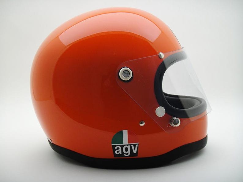 May include: Glossy orange motorcycle helmet with a clear visor and black trim. The helmet has a small green, white, and black logo on the side. A classic design for motorcycle enthusiasts.