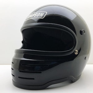 May include: A glossy black full-face motorcycle helmet featuring a prominent Simpson brand logo in white text on the forehead area. The helmet includes a wide eye port, a chin bar with three horizontal ventilation slits, and silver hardware.