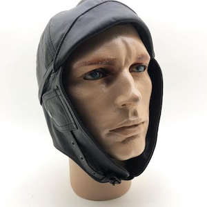 Può includere: Cappello da aviatore in pelle nera con paraorecchie e visiera. Il cappello ha una chiusura a bottone sui paraorecchie e un cinturino sotto il mento. Il cappello è esposto su una testa di manichino.
