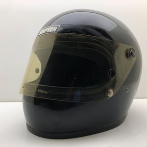 Op de afbeelding: Een glanzende zwarte integraalhelm voor motorrijders met de merknaam SIMPSON in witte letters op het voorhoofd. De helm is voorzien van een getint, halfdoorzichtig vizier met zichtbare bevestigingsonderdelen aan de zijkanten en een helder lensgedeelte.