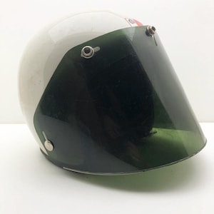 May include: A vintage white motorcycle helmet with a dark green visor. The helmet has a glossy finish and a red and white stripe detail. The visor is attached with metal hardware. This helmet is designed for safety and protection.
