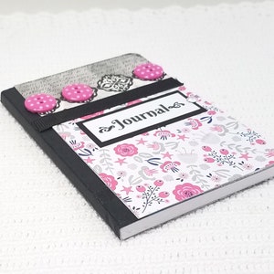 May include: A small black journal with a floral design and pink buttons. The journal has a black ribbon and the word "Journal" written on the front.