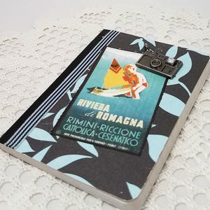 Puede incluir: Una libreta negra y azul con un cartel de viaje vintage que muestra un velero y el texto "Riviera di Romagna Rimini-Riccione Cattolica-Cesenatico". Un charm de c&aacute;mara plateado cuelga de la libreta.