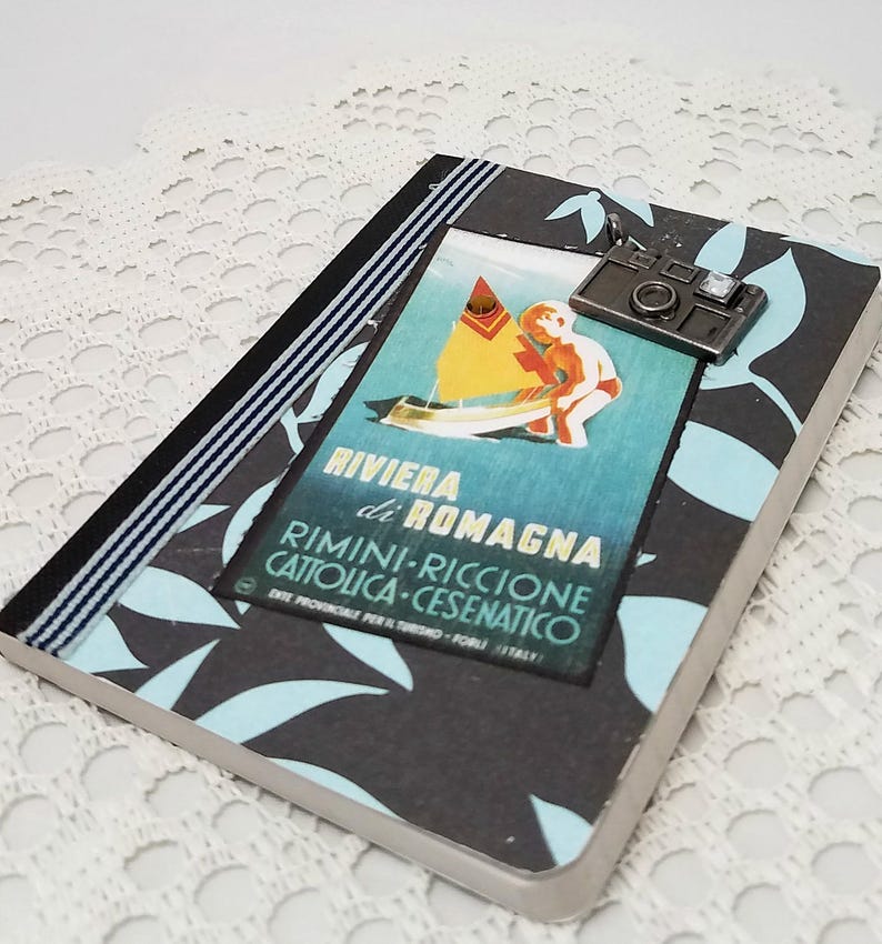 Puede incluir: Una libreta negra y azul con un cartel de viaje vintage que muestra un velero y el texto "Riviera di Romagna Rimini-Riccione Cattolica-Cesenatico". Un charm de c&aacute;mara plateado cuelga de la libreta.