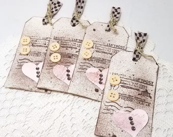 Puede incluir: Cuatro etiquetas de regalo de papel marrón con un corazón rosa, un sello vintage que dice "Deutsche Luftpost" y tres botones. Las etiquetas están atadas con una cinta a cuadros marrón y blanca.