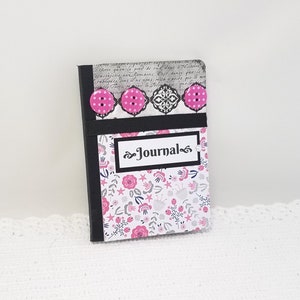 May include: A small black journal with a floral design and pink buttons. The cover has a black label with the word "Journal" written in white.