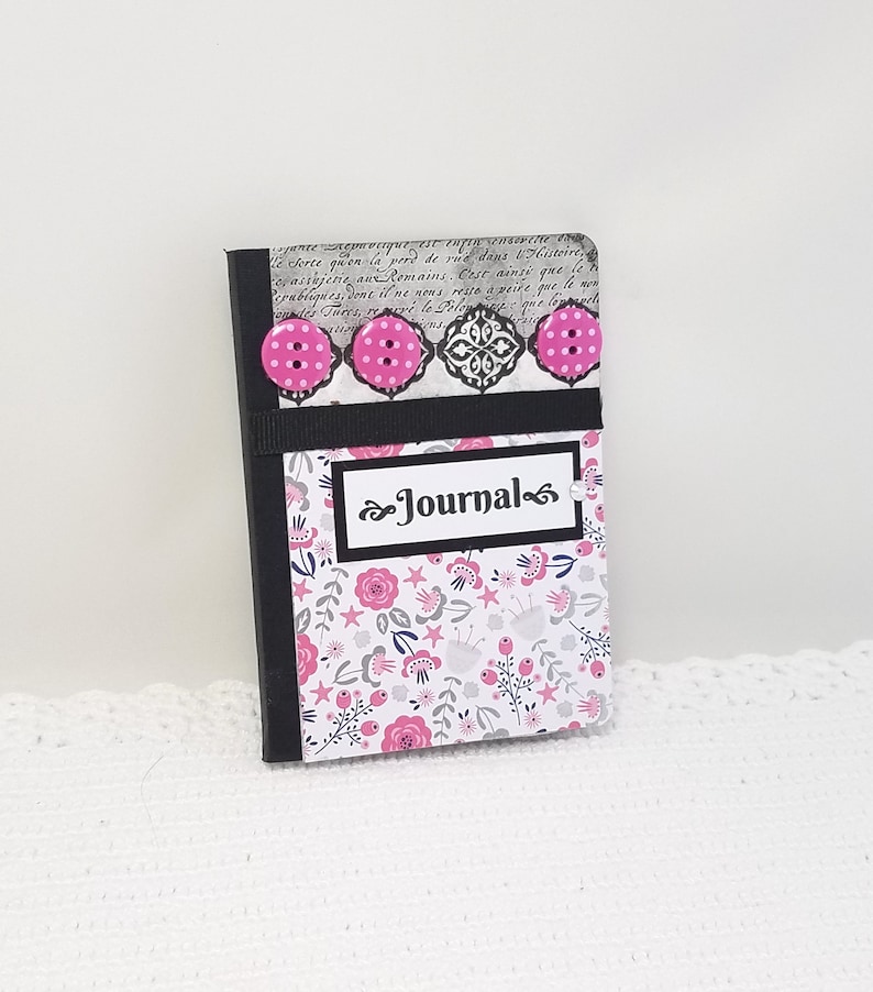 May include: A small black journal with a floral design and pink buttons. The cover has a black label with the word "Journal" written in white.