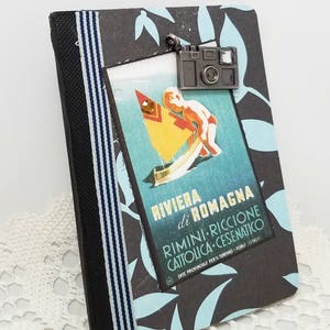 Puede incluir: Un diario a rayas negras y azules con un cartel de viaje vintage en la parte delantera. El cartel presenta a un ni&ntilde;o en un velero con el texto "Riviera di Romagna Rimini-Riccione Cattolica-Cesenatico Ente Provinciale per il Turismo - Forli - Italy". Un amuleto de c&aacute;mara plateado cuelga de la parte superior del diario.