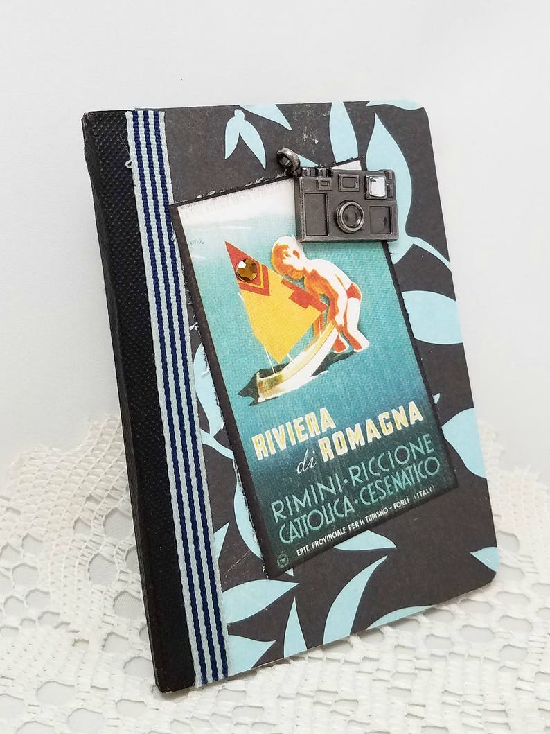 Puede incluir: Un diario a rayas negras y azules con un cartel de viaje vintage en la parte delantera. El cartel presenta a un ni&ntilde;o en un velero con el texto "Riviera di Romagna Rimini-Riccione Cattolica-Cesenatico Ente Provinciale per il Turismo - Forli - Italy". Un amuleto de c&aacute;mara plateado cuelga de la parte superior del diario.