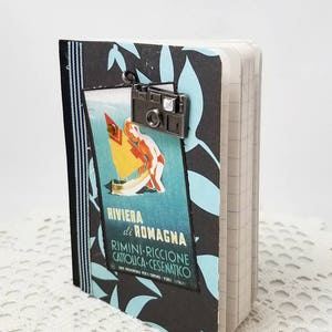 Puede incluir: Una libreta negra y azul con un cartel de viaje vintage en la portada. El cartel presenta un velero azul y blanco y el texto "RIVIERA di ROMAGNA RIMINI-RICCIONE CATTOLICA-CESENATICO". Un peque&ntilde;o charm de c&aacute;mara plateado est&aacute; unido a la parte superior de la libreta.