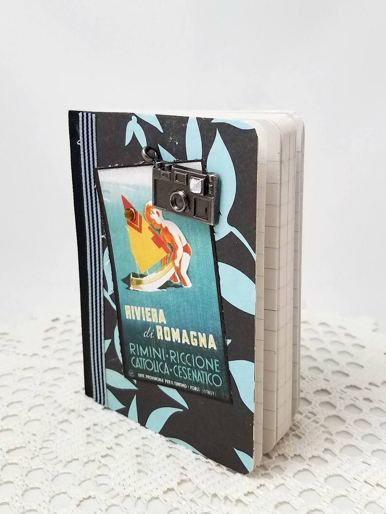 Puede incluir: Una libreta negra y azul con un cartel de viaje vintage en la portada. El cartel presenta un velero azul y blanco y el texto "RIVIERA di ROMAGNA RIMINI-RICCIONE CATTOLICA-CESENATICO". Un peque&ntilde;o charm de c&aacute;mara plateado est&aacute; unido a la parte superior de la libreta.