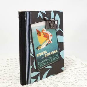 Puede incluir: Un diario de viaje negro y azul con un cartel de viaje vintage en la parte delantera. El cartel presenta a un ni&ntilde;o en un velero con el texto "Riviera di Romagna Rimini-Riccione Cattolica-Cesenaico". Un amuleto de c&aacute;mara plateado cuelga de la parte superior del diario.