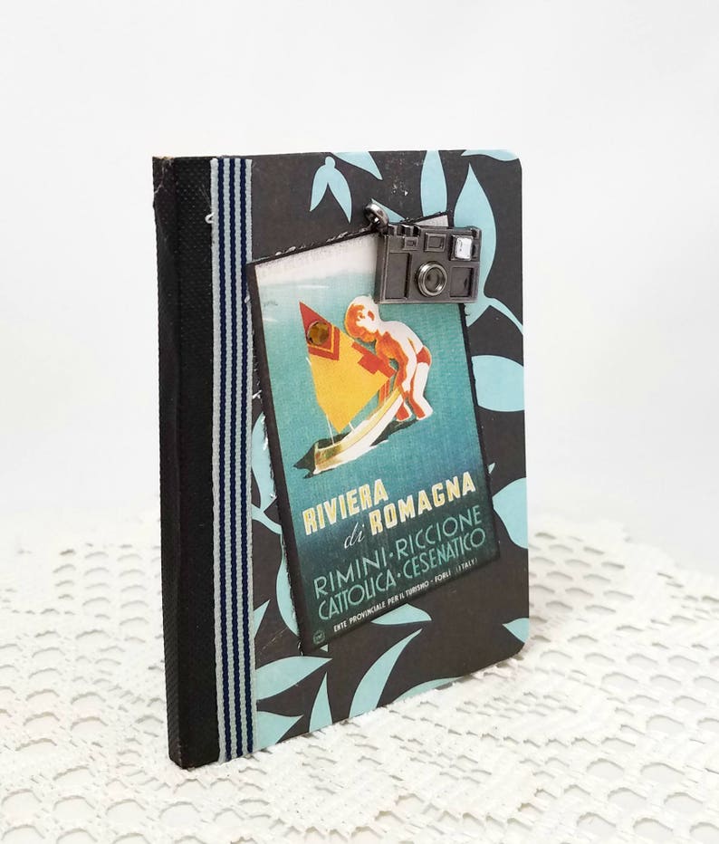 Puede incluir: Un diario de viaje negro y azul con un cartel de viaje vintage en la parte delantera. El cartel presenta a un ni&ntilde;o en un velero con el texto "Riviera di Romagna Rimini-Riccione Cattolica-Cesenaico". Un amuleto de c&aacute;mara plateado cuelga de la parte superior del diario.