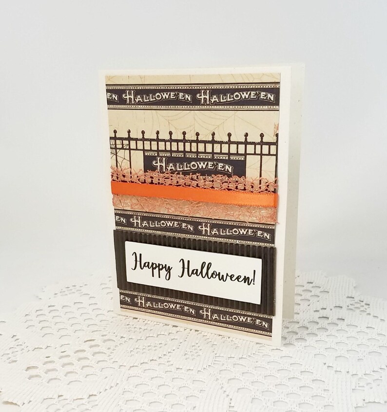 May include: A Halloween greeting card with a decorative fence design and the word "Halloween" repeated. The card features orange ribbon and the message "Happy Halloween!" on a white rectangle.