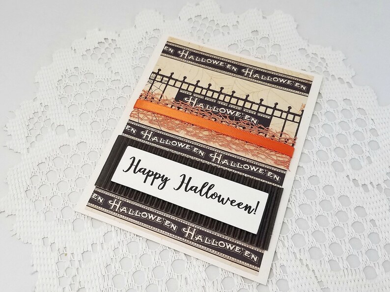 May include: A Halloween greeting card with a decorative fence design, orange ribbon, and the words "Happy Halloween!" in a white rectangle. The card has a vintage aesthetic with a cream and black color scheme.