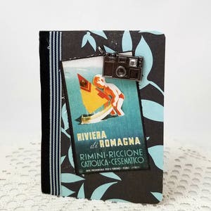 Puede incluir: Un diario de viaje negro y azul con un amuleto de c&aacute;mara vintage. La portada presenta un cartel de viaje vintage con el texto "Riviera di Romagna Rimini-Riccione Cattolica-Cesenatico."