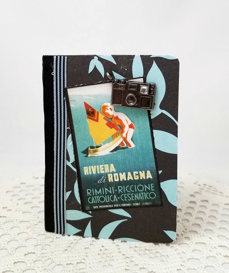 Puede incluir: Un diario de viaje negro y azul con un amuleto de c&aacute;mara vintage. La portada presenta un cartel de viaje vintage con el texto "Riviera di Romagna Rimini-Riccione Cattolica-Cesenatico."