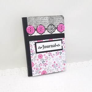 May include: A small pink and black journal with a floral pattern and the word "Journal" on the front cover. The journal has three pink buttons with white polka dots.