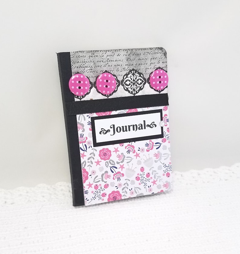 May include: A small pink and black journal with a floral pattern and the word "Journal" on the front cover. The journal has three pink buttons with white polka dots.