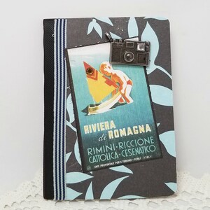 Puede incluir: Una libreta de rayas negras y azules con un cartel de viaje vintage en la portada. El cartel muestra a un ni&ntilde;o en un velero con el texto "Riviera di Romagna Rimini-Riccione Cattolica-Cesenatico Ente Provinciale per il Turismo - Forli Italy". Un charm de c&aacute;mara plateado est&aacute; unido a la libreta.