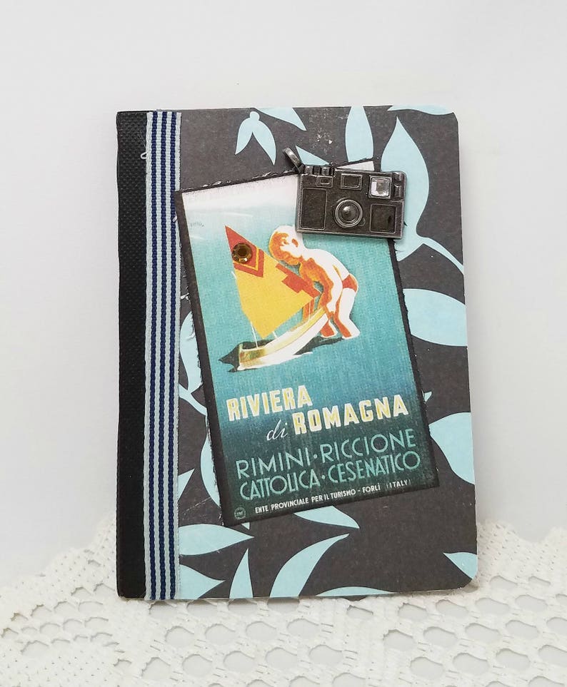 Puede incluir: Una libreta de rayas negras y azules con un cartel de viaje vintage en la portada. El cartel muestra a un ni&ntilde;o en un velero con el texto "Riviera di Romagna Rimini-Riccione Cattolica-Cesenatico Ente Provinciale per il Turismo - Forli Italy". Un charm de c&aacute;mara plateado est&aacute; unido a la libreta.