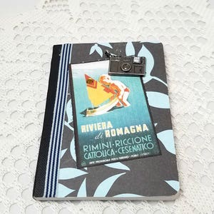 Puede incluir: Una libreta negra y azul con un cartel de viaje vintage en la parte delantera. El cartel muestra a un ni&ntilde;o en un velero con el texto "Riviera di Romagna" y "Rimini-Riccione Cattolica-Cesenatico". Un charm de c&aacute;mara plateado cuelga de la parte superior de la libreta.