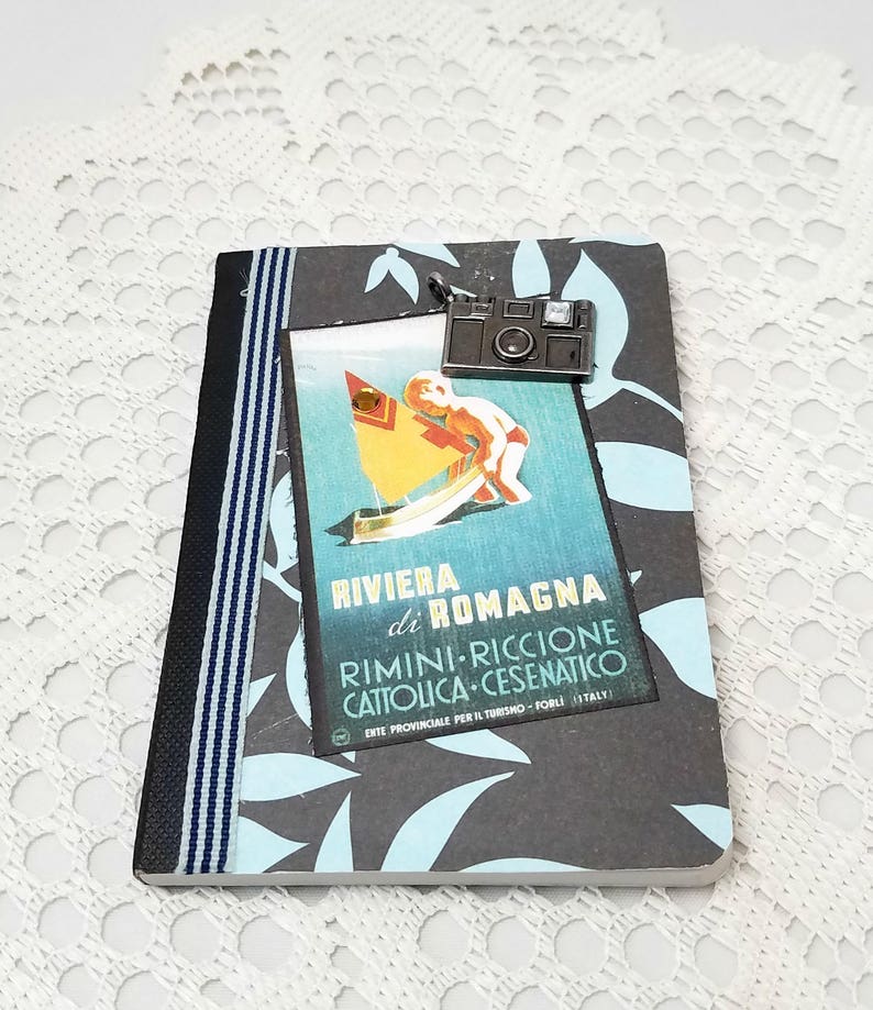 Puede incluir: Una libreta negra y azul con un cartel de viaje vintage en la parte delantera. El cartel muestra a un ni&ntilde;o en un velero con el texto "Riviera di Romagna" y "Rimini-Riccione Cattolica-Cesenatico". Un charm de c&aacute;mara plateado cuelga de la parte superior de la libreta.