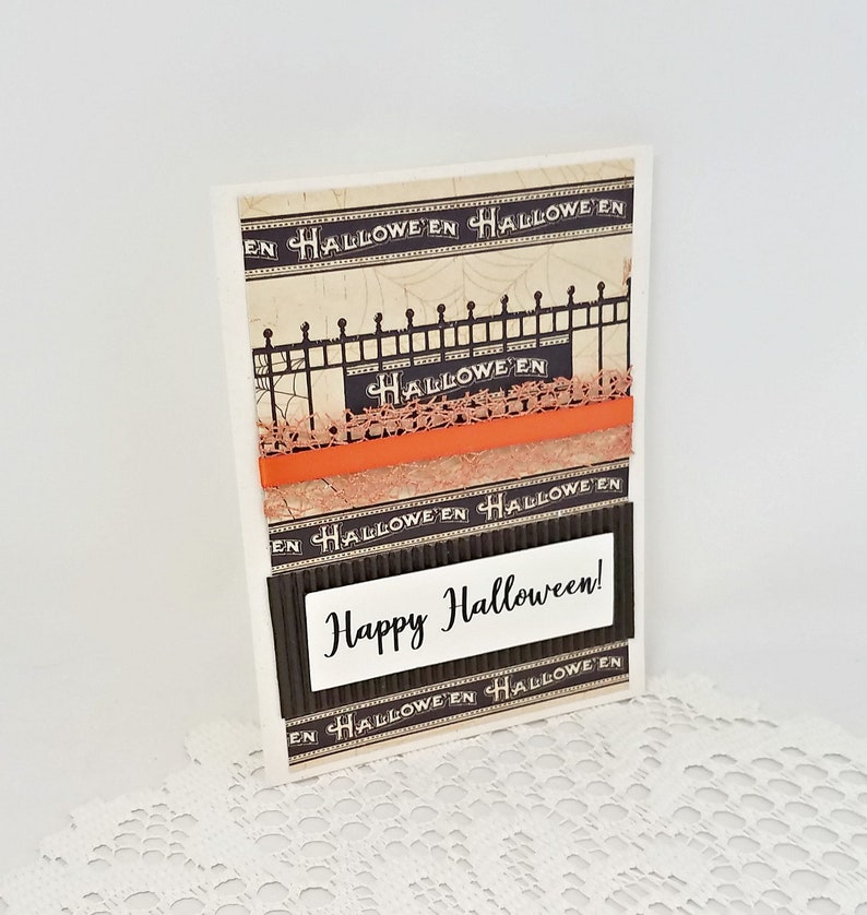 May include: A handmade Halloween greeting card with a vintage aesthetic. The card features a black fence design, orange accents, and the words "Happy Halloween!" in a white rectangle. The background has a repeating "Halloween" pattern.