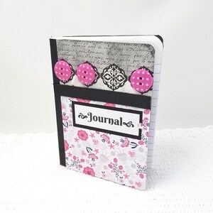 May include: A small pink and white journal with a black ribbon and pink buttons. The cover has a floral pattern and the word "Journal" is written in black cursive.
