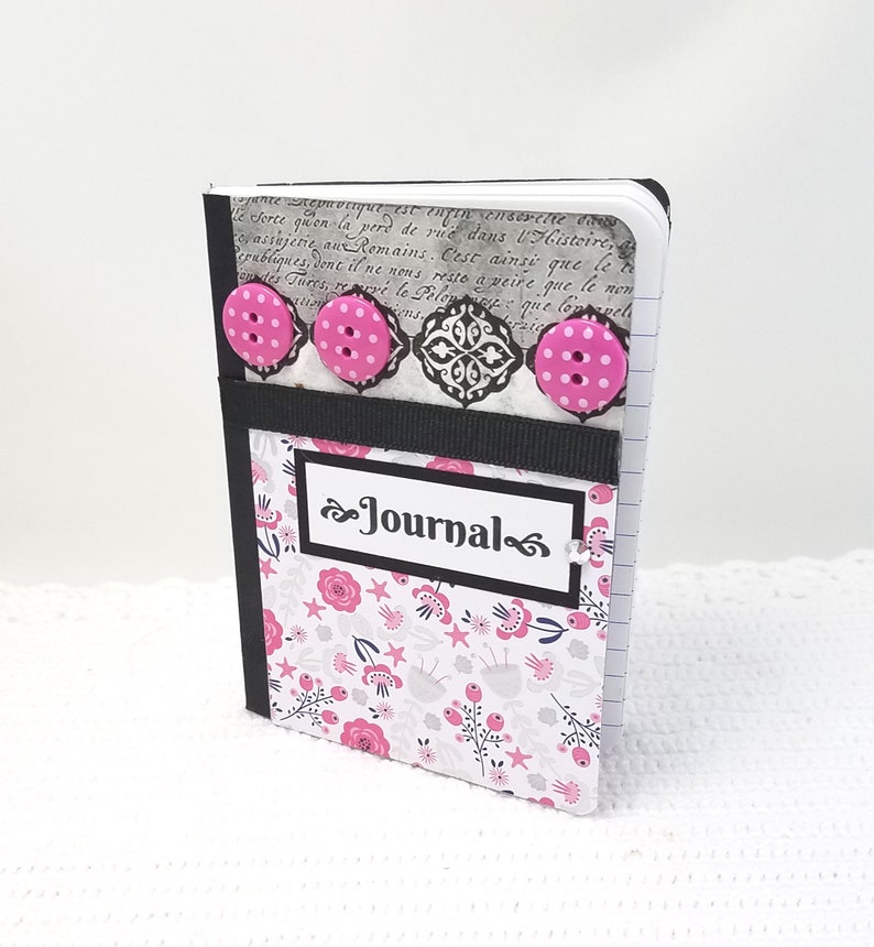 May include: A small pink and white journal with a black ribbon and pink buttons. The cover has a floral pattern and the word "Journal" is written in black cursive.