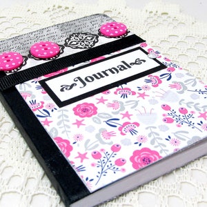 May include: A black journal with a floral design and pink buttons. The cover has a black ribbon and a white label with the word "Journal" written in black cursive.