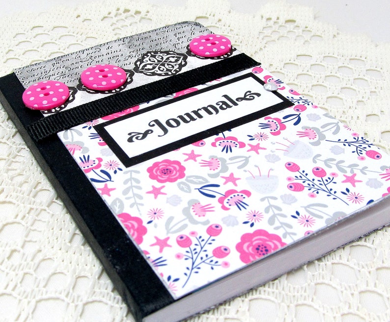 May include: A black journal with a floral design and pink buttons. The cover has a black ribbon and a white label with the word "Journal" written in black cursive.