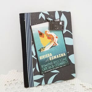 Puede incluir: Un peque&ntilde;o cuaderno gris decorado con una cinta a rayas azul y negra. Un p&oacute;ster vintage de viaje con un ni&ntilde;o en un velero est&aacute; pegado en el cuaderno. El p&oacute;ster dice "Riviera di Romagna Rimini - Riccione Cattolica - Cesenatico Ente Provinciale per il Turismo - Forli - Italy". Un amuleto de c&aacute;mara plateado cuelga de la parte superior del cuaderno.