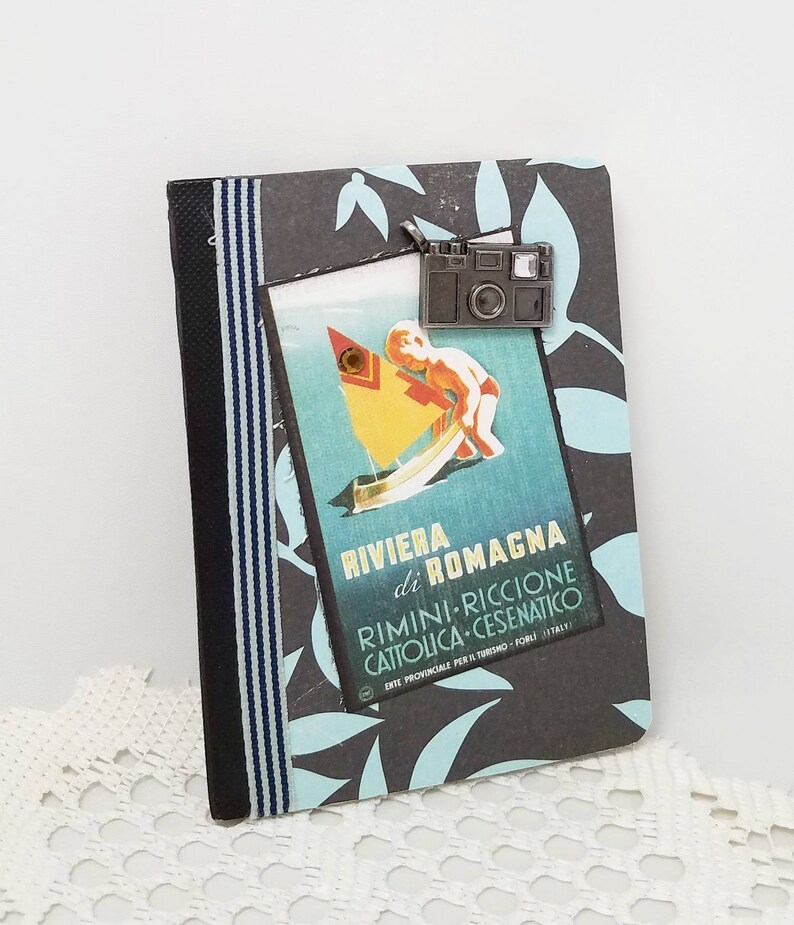 Puede incluir: Un peque&ntilde;o cuaderno gris decorado con una cinta a rayas azul y negra. Un p&oacute;ster vintage de viaje con un ni&ntilde;o en un velero est&aacute; pegado en el cuaderno. El p&oacute;ster dice "Riviera di Romagna Rimini - Riccione Cattolica - Cesenatico Ente Provinciale per il Turismo - Forli - Italy". Un amuleto de c&aacute;mara plateado cuelga de la parte superior del cuaderno.