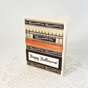 May include: A handmade Halloween greeting card with a cream-colored background. The card features decorative black and orange ribbons, a black fence design, and the words "Happy Halloween!" in white text.
